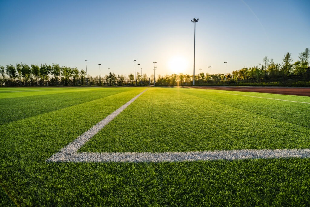 A vibrant, sunlit soccer field with neatly marked white lines, surrounded by tall light poles and trees at the horizon, under a clear sky.