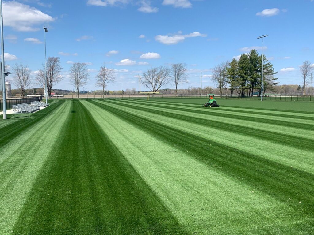 A person mows a neatly striped sports field, surrounded by trees and benches, under a clear blue sky with scattered clouds.