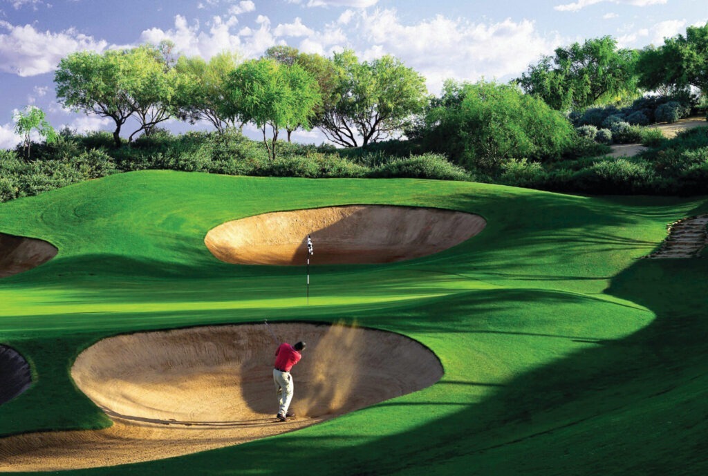 A person in red shirt plays golf from a bunker on a lush green course, surrounded by trees under a partly cloudy sky.