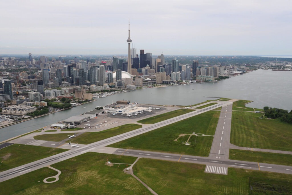 The image shows an aerial view of Toronto with the CN Tower, surrounded by high-rise buildings, and an adjacent airport runway near Lake Ontario.