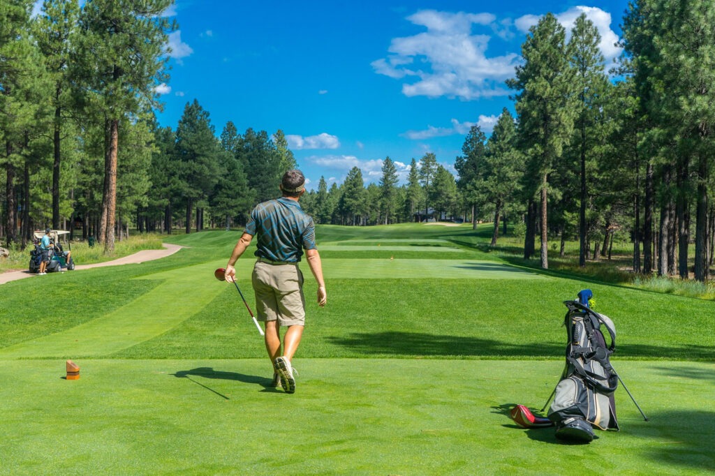 A person on a vibrant green golf course, surrounded by tall trees, walks away with clubs. Another person drives a golf cart nearby.