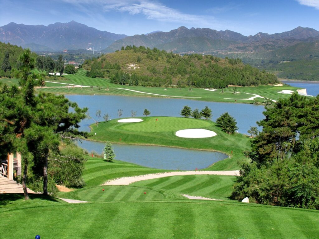 A scenic golf course featuring lush greens, water hazards, and mountainous backdrops, surrounded by pine trees on a clear day.