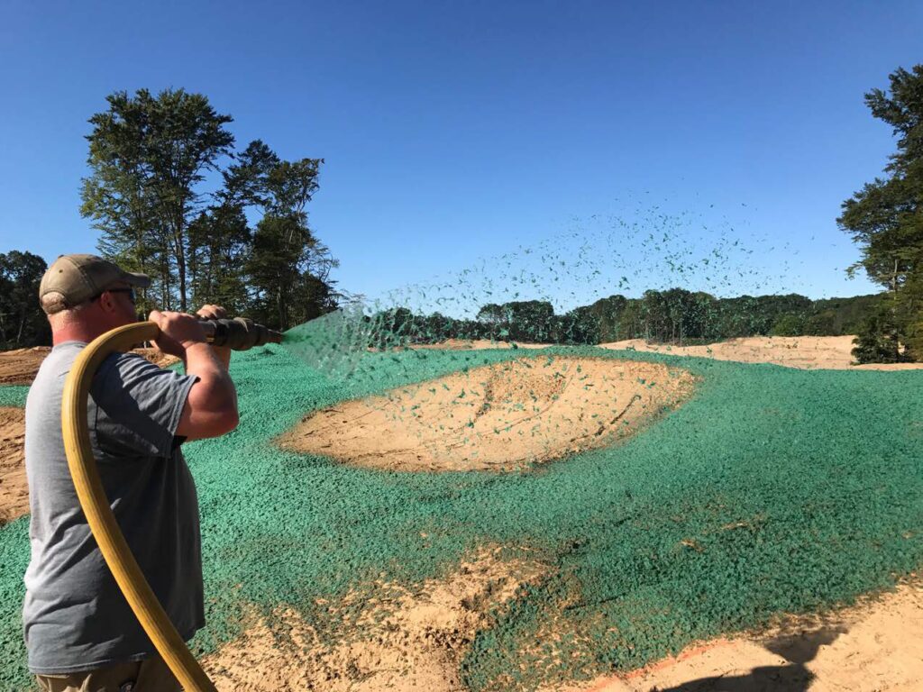 A person sprays green hydroseed onto a dry landscape with trees in the background under a clear blue sky.