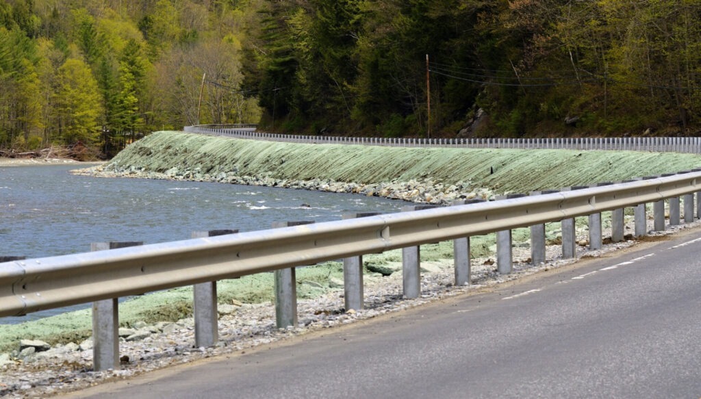 A winding road with metal guardrails runs alongside a river, surrounded by a forested area with lush green trees and a rocky embankment.