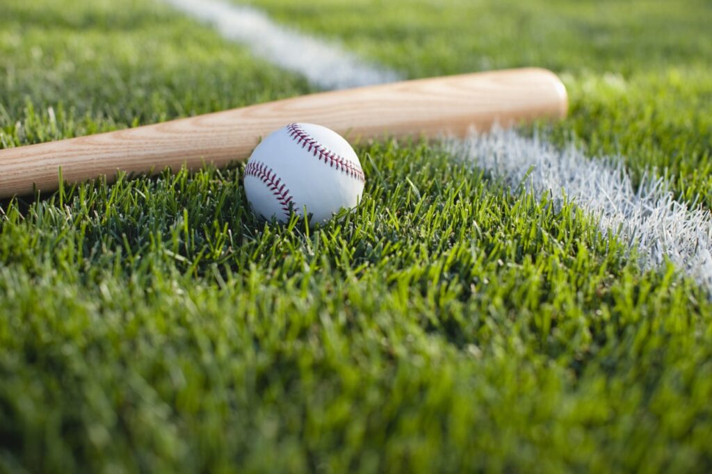 A baseball and bat resting on freshly cut grass near a white chalk line on a baseball field, under clear daylight conditions.