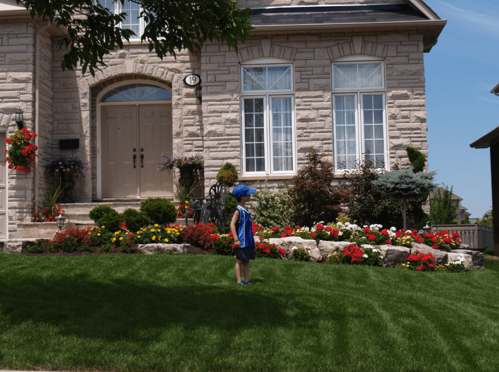 A child wearing a blue outfit stands on the lawn of a stone house with a well-maintained garden full of colorful flowers.