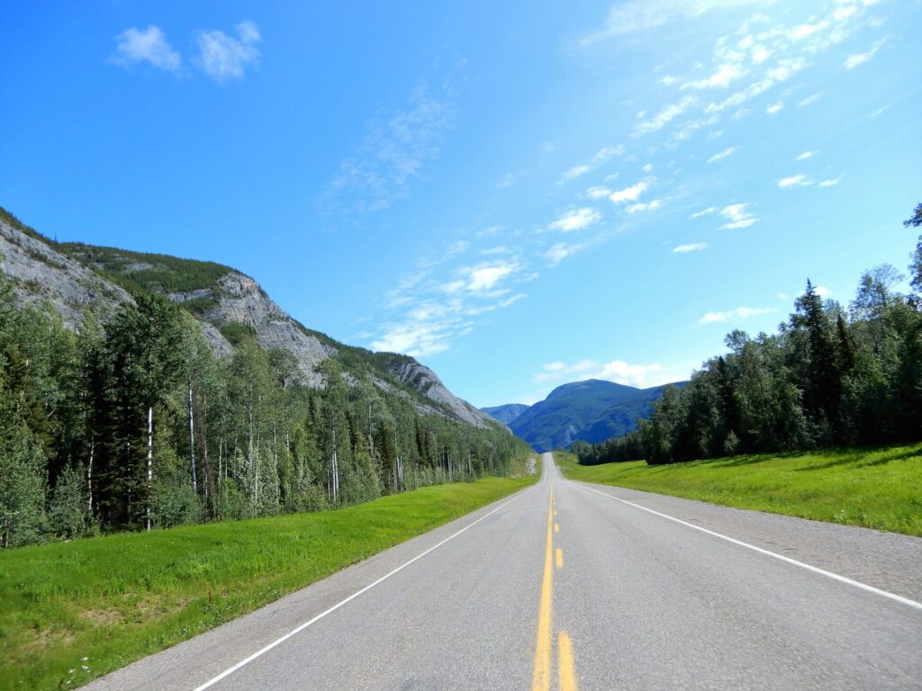 A straight road winds through a mountainous landscape with green forests on either side, under a clear, bright blue sky.