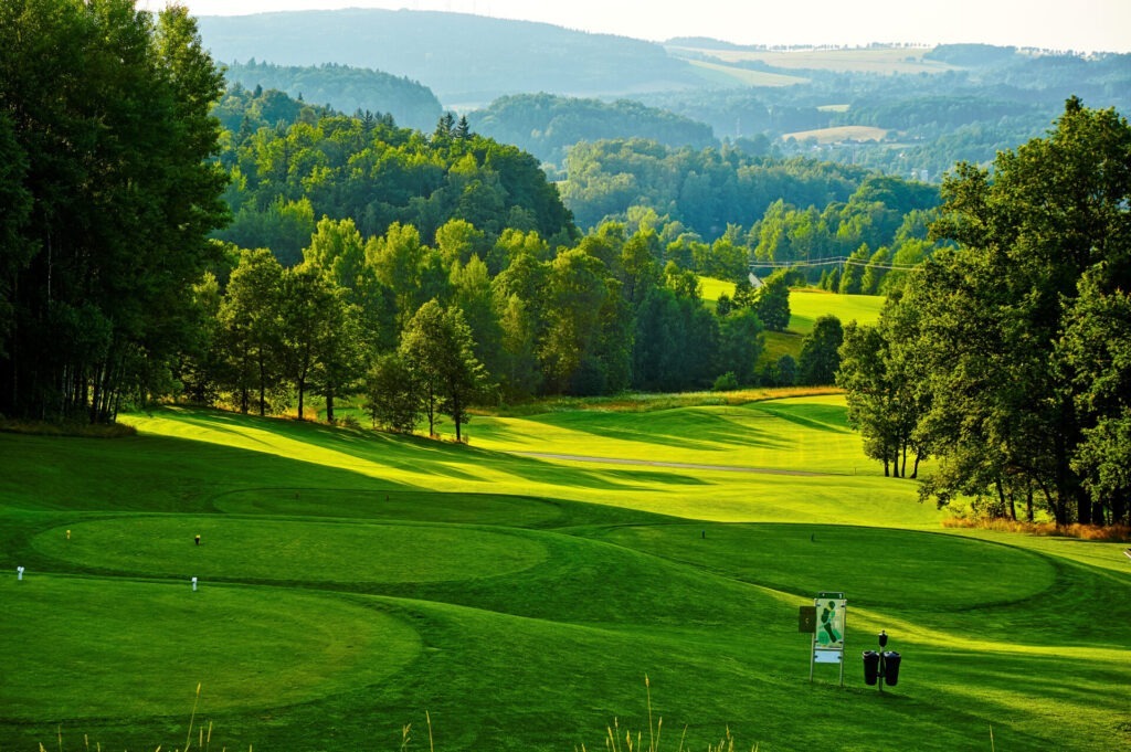 A lush, green golf course surrounded by trees and hills, with a person in the distance near a golf bag under a clear sky.