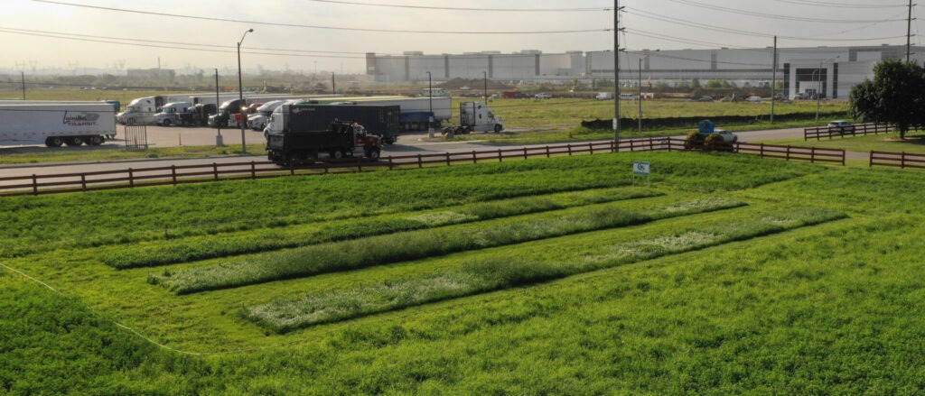 A grassy field with vehicular traffic in the background, divided by fences. Trucks are parked near a large warehouse complex on the horizon.