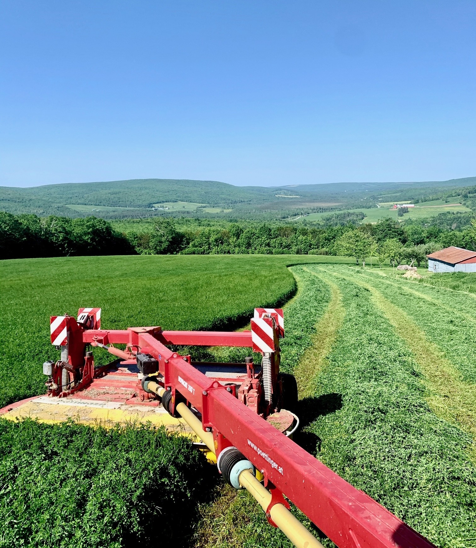 A red agricultural machine cuts through a lush green field with rolling hills and a blue sky in the background.