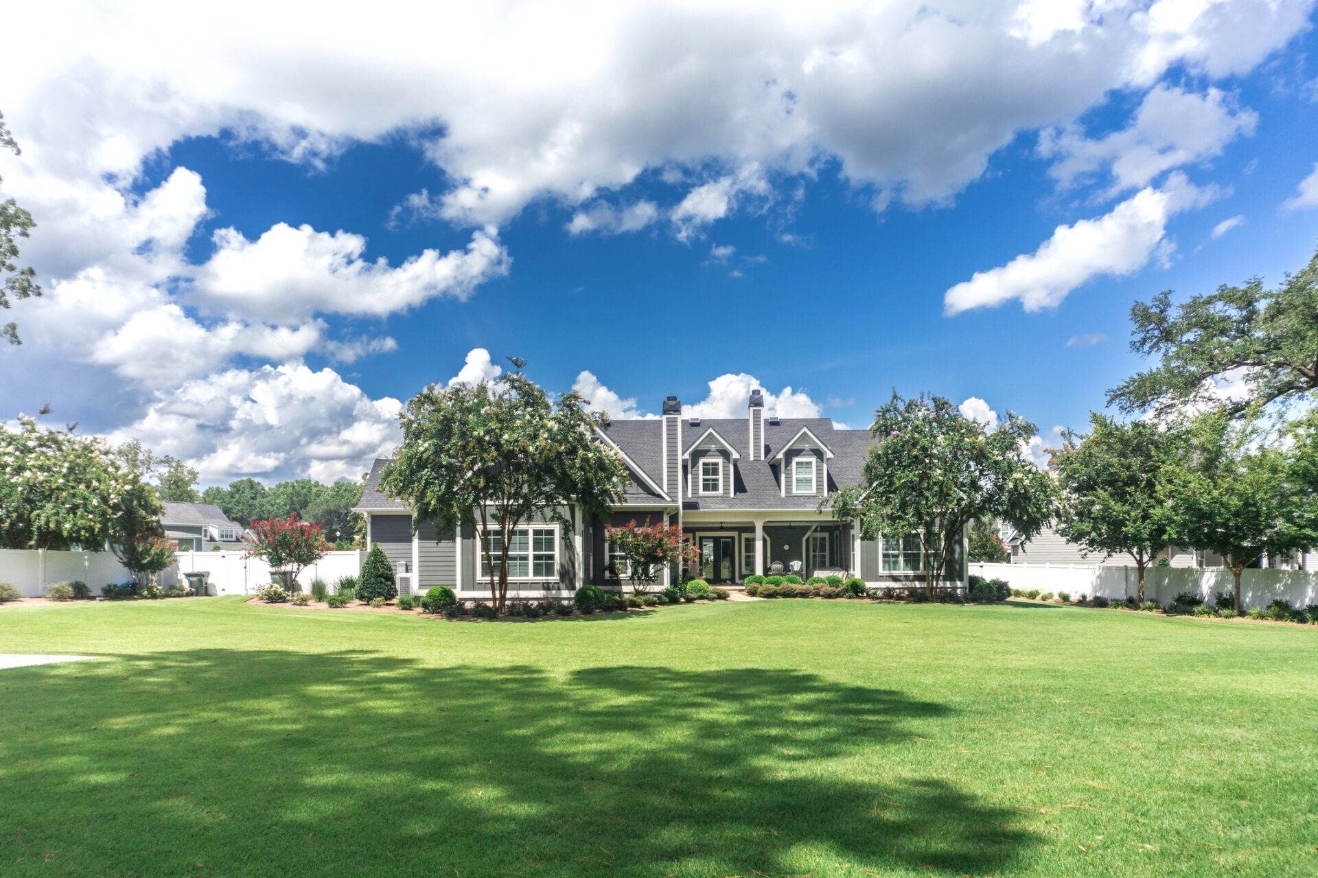 A large house with a well-maintained green lawn, manicured shrubs, and trees under a bright, blue sky with scattered clouds. No recognizable landmarks.