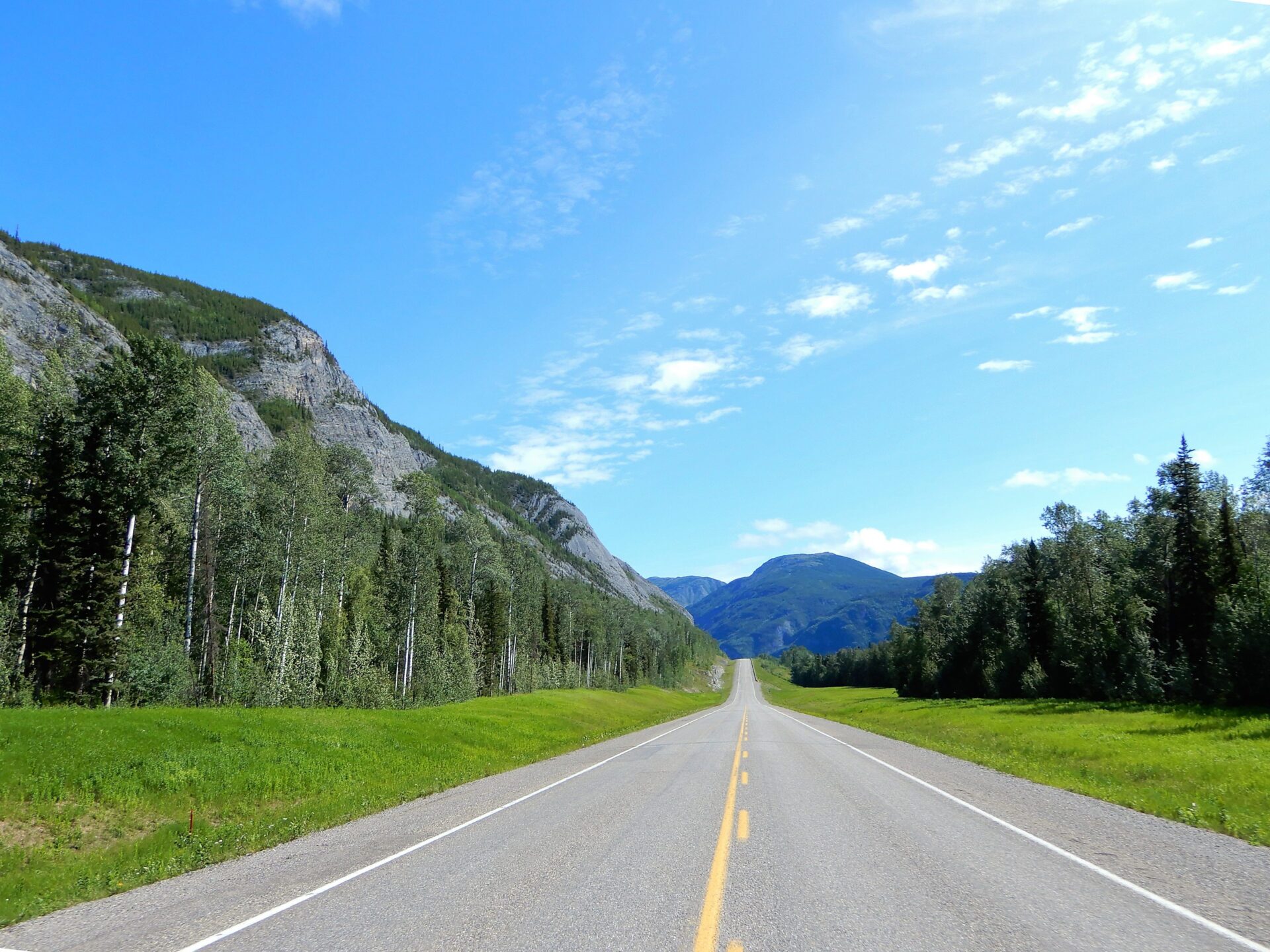 A long, empty road stretches through a mountainous landscape filled with lush green trees under a bright blue sky with scattered clouds.