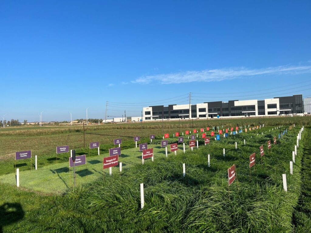 The image shows a grassy field with labeled plant plots, situated in front of a modern industrial building under a clear blue sky.