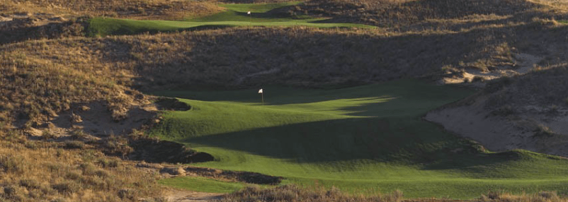 A tranquil golf course with rolling hills, neatly trimmed greens, and a visible flagstick under gentle sunlight, surrounded by dry, brushy vegetation.