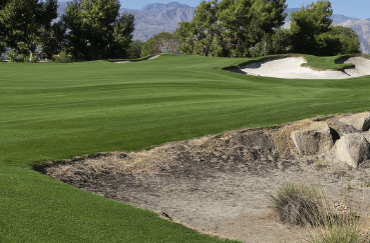 Lush green golf course with sand bunkers, trees, and distant mountains under a clear sky. No landmarks or historical buildings visible.