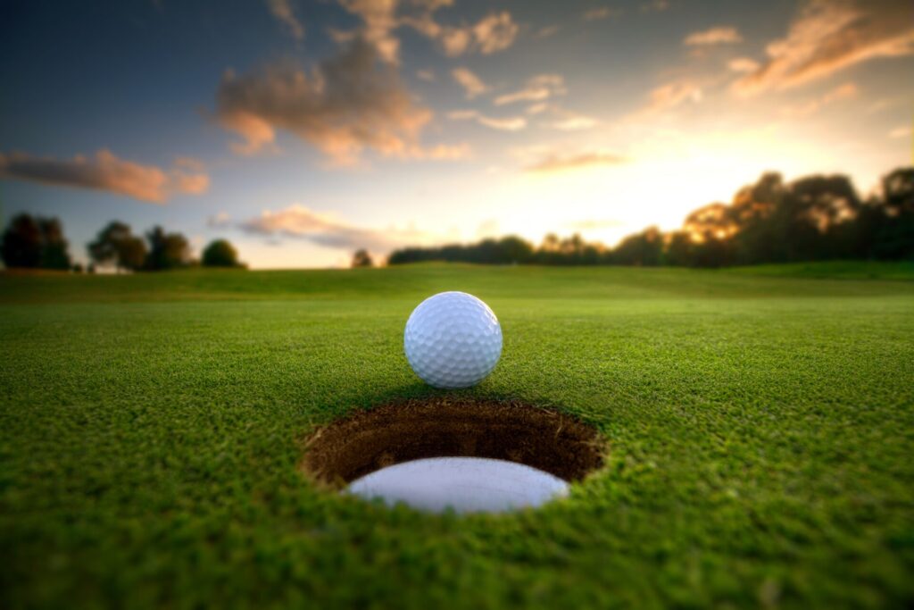 A golf ball sits at the edge of a hole on a well-manicured green, with a scenic sunset and trees in the background.