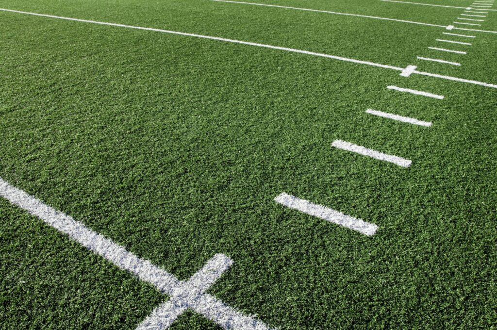 A close-up view of a green artificial turf football field with white yard lines clearly marked, no people or landmarks visible.