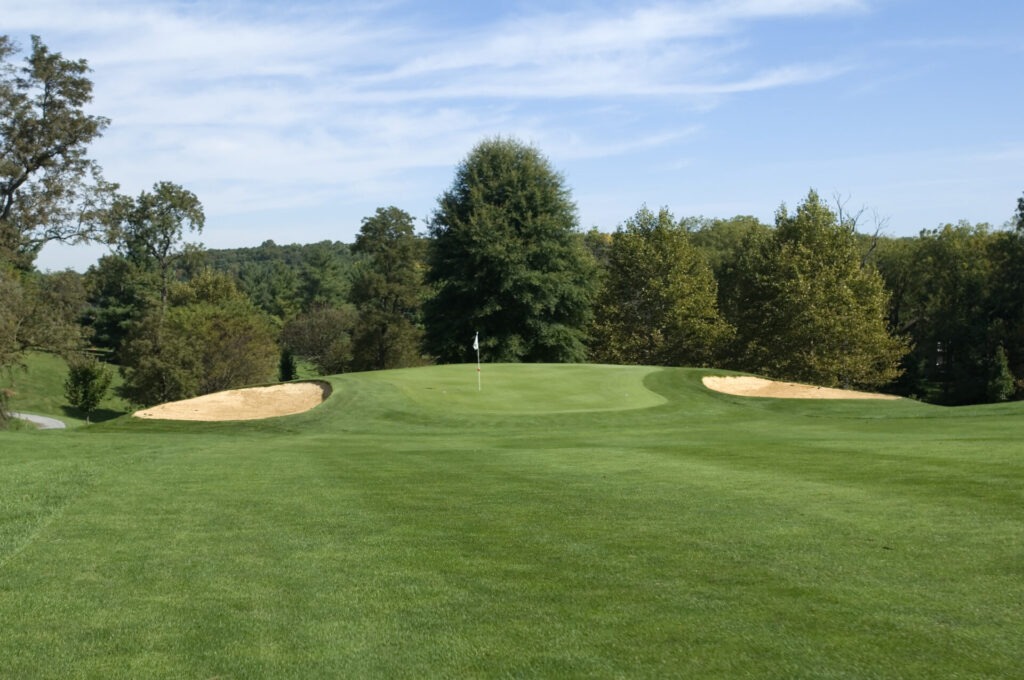 A lush, green golf course with sand traps, a flag, and surrounding trees under a partly cloudy sky.