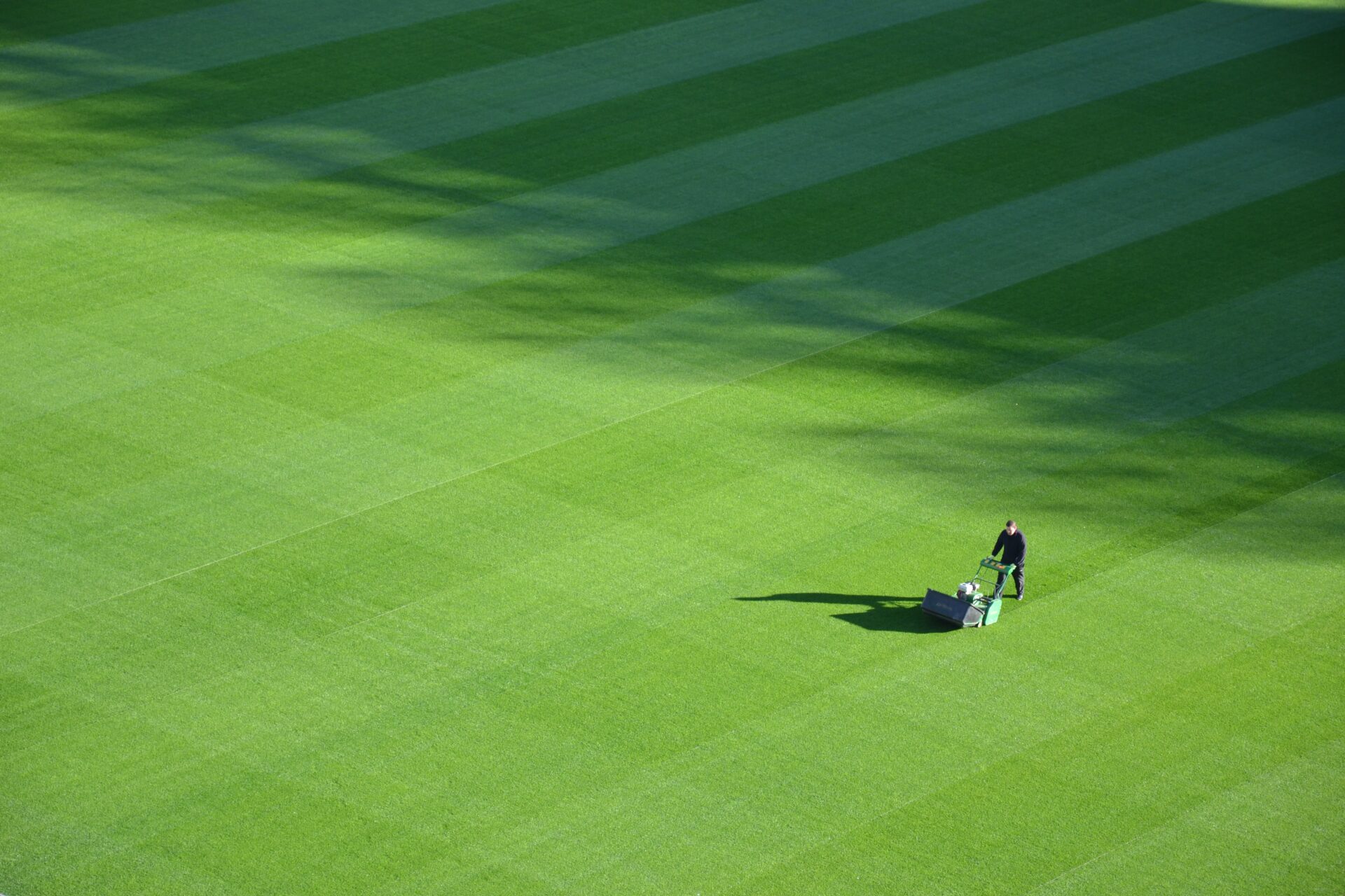 A person mowing green grass on a large, striped lawn, casting a long shadow under clear daylight with no recognizable landmarks visible.