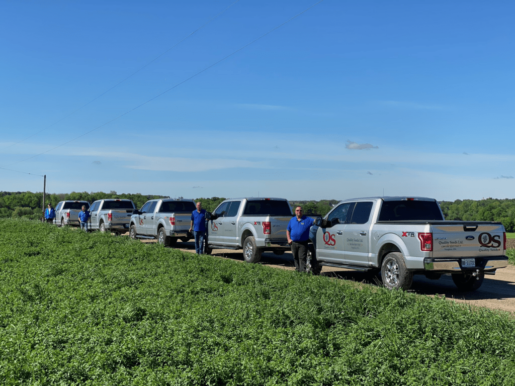 Several persons in blue shirts stand beside silver trucks marked with "Quality Seeds Ltd." on a rural road, with greenery and clear skies in the background.